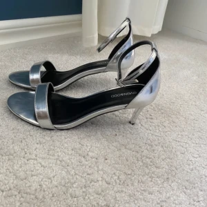 Silvriga klackar Even&Odd - Snygga sandaler i metallic silver från Even&Odd med smal rem över tårna och justerbar ankelrem. Skorna har öppen tå och smal klack som ger en elegant look. Perfekta för dig som vill sticka ut med stilrena detaljer.