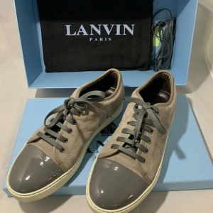 Grå sneakers från Lanvin med mocka - Snygga sneakers från Lanvin i grå mocka med matchande grå snörning och glansig grå tå i syntet. Skorna har en vit sula och klassisk låg profil. Perfekta för dig som gillar stilrena och exklusiva detaljer.
