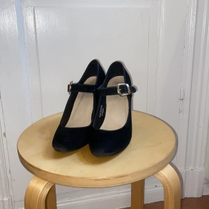 Svarta Mary Jane pumps från mrp - Klassiska svarta Mary Jane pumps med rund tå och bred klack. Skorna har en snygg rem över foten med guldfärgat spänne och är gjorda i mjuk mocka-look. Perfekta för dig som gillar retrovibbar och vill ha en bekväm men stilren klack.
