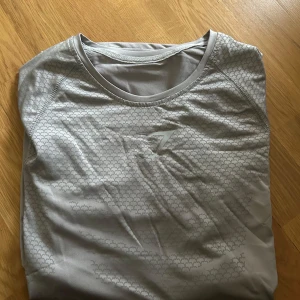 Ljusgrå Gymshark t-shirt med mönster - Snygg ljusgrå t-shirt från Gymshark med diskret geometriskt mönster över hela tröjan och rund halsringning. Perfekt för träning med sitt lätta och stretchiga material. Gymshark-logga på bröstet ger en sportig vibe.