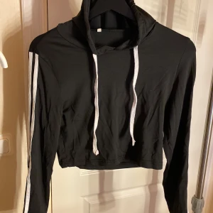 Svart croppad hoodie med vita ränder - Svart croppad hoodie med långa ärmar och vita ränder längs ärmarna. Tröjan har huva och vita snören framtill för en sportig vibe. 