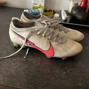 Nike Mercurial fotbollsskor vit/röd - Nike Mercurial fotbollsskor i vit med stora röda och turkosa swoosh-loggor. Skorna har snörning, platt sula med dobbar i rött och blått, och är tillverkade i syntetmaterial med textilstruktur. Perfekta för dig som vill sticka ut på planen.