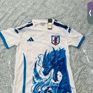 Japan fotbollströja Adidas blå drake - Japan landslagets fotbollströja från Adidas med en stor blå drake på framsidan och blå detaljer på ärmar och axlar. Tröjan är vit med blått tryck, har korta ärmar och officiellt klubbmärke på bröstet. Perfekt för dig som vill sticka ut på matchen.
