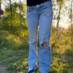 Lågmidjade ljusblå bootcut jeans - Säljer ett par lågmidjade ljusblå jeans från True Religion med bootcut passform. Jeansen har slitna detaljer och hål på låret samt bakfickor med broderade detaljer och knappar. Perfekta för dig som gillar en avslappnad och trendig look!!! 👀💞pris kan diskuteras vid snabb affär!