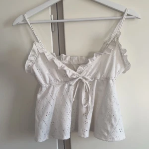 Vit broderad linnetopp med volang - Supersöt vit linnetopp med broderade hålmönster, smala axelband och volangdetaljer längs urringningen. Toppen har knyt framtill och är lätt utsvängd för en luftig känsla. Perfekt för varma dagar.💞