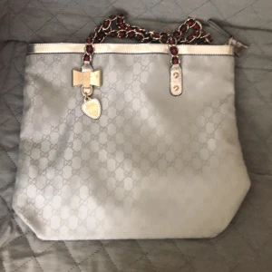 Gucci axelväska med kedjedetaljer - Snygg axelväska från Gucci i ljusgrått med klassiskt GG-mönster. Väskan har guldfärgade detaljer, en dekorativ rosett och hjärtberlock framtill samt kedjerem med inslag av rött och svart. Insidan har ett unikt mönster i beige, brunt och orange.