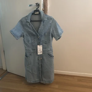 Ljusblå jeansklänning med fickor - Snygg ljusblå jeansklänning från Zara med kort ärm, knappar framtill och fyra stora fickor. Klassisk skjortkrage och avslappnad passform, perfekt för en cool och avslappnad look. Materialet är jeans och klänningen slutar ovanför knäna. Kan  passa  Stl. S 