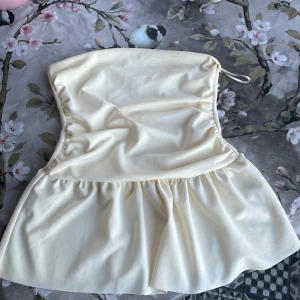 Krämvit bandeau tubtopp Gina Tricot - Supersöt bandeau tubtopp från Gina Tricot i krämvit färg. Toppen är axelbandslös med volangliknande nederdel och har en dragsko upptill för perfekt passform. Perfekt för sommarens alla tillfällen och lätt att styla med jeans eller kjol.