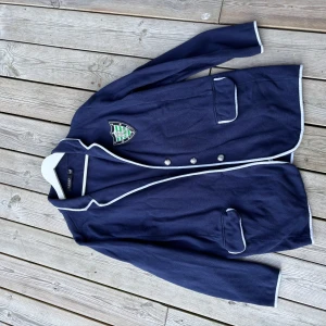 Marinblå kavaj från Lauren Ralph Lauren - Snygg marinblå kavaj från Lauren Ralph Lauren med vita kanter, emblem på bröstet och dubbla fickor framtill. Klassisk design med knappar och lång ärm, perfekt för en preppy look. Materialet är mjukt och känns lyxigt.