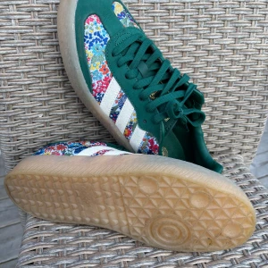 Adidas Samba blommiga sneakers gröna - Unika Adidas Samba sneakers med gröna detaljer och blommigt mönster i flera färger. Klassiska vita ränder på sidorna och gummisula. Perfekta för dig som vill sticka ut med färg och stil. Skorna har snörning och rund tå.