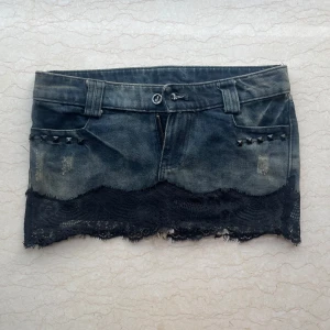 Jeanskjol med spets och nitar - Cool kort jeanskjol i mörkblå denim med slitna detaljer, svarta nitar och bred svart spetskant längst ner. Kjolen har bälteshällor, fickor fram och bak samt dragkedja. Perfekt för dig som gillar edgy och unik stil.