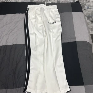 Vita Palm Angels trackpants med svarta ränder - Palm Angels vita trackpants med svarta sidoränder och broderad logga framtill. Byxorna har elastisk midja med snörning och dragkedjor vid bensluten. Tillverkade i ett glansigt syntetmaterial som ger en sportig vibe.