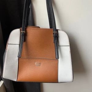 Guess handväska  - Snygg handväska från Guess i brunt och vitt med svarta detaljer. Väskan har flera fack, dragkedja och magnetlås. Insidan är fodrad med logotypmönster och har både öppna och stängbara fack. Perfekt storlek för att få plats med allt du behöver.🌸