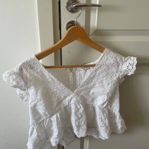 Vit broderad blus från NA-KD - Superfin vit blus från NA-KD i bomull med broderade detaljer och volangkant. Blusen har v-ringning, korta ärmar med spets och en luftig, croppad passform. Perfekt för sommarens alla tillfällen och ger en romantisk vibe.