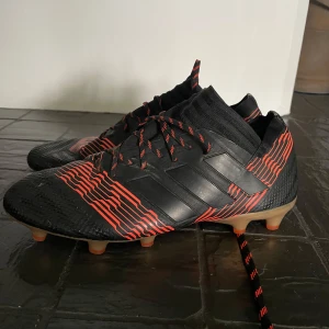 Adidas svarta och röda fotbollsskor - Snygga fotbollsskor från Adidas i svart med röda linjer och matchande snören. Skorna har en strumpliknande passform och plös, samt dobbar för spel på gräs. Tillverkade i syntetmaterial för bästa grepp och känsla på planen.
