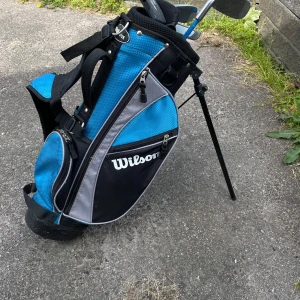 Wilson blå golfbag med stativ - Säljer en sportig golfbag från Wilson i blått, svart och grått med praktiskt stativ och flera fack med dragkedja. Väskan har vadderade axelremmar och är tillverkad i slitstark polyester. Perfekt för att bära golfklubbor på banan.