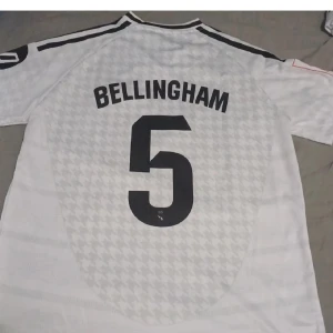 Real Madrid Bellingham 5 vit matchtröja - Snygg Real Madrid fotbollströja med Bellingham och nummer 5 på ryggen. Vit med subtilt rutigt mönster, svarta detaljer och klubbmärke på bröstet. Adidas-logga och sponsortryck framtill. Kortärmad och tillverkad i lätt funktionsmaterial.