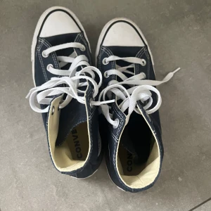 Mörkblå Converse All Star high tops - Klassiska mörkblå Converse All Star high tops med vit sula och vita snören. Skorna har rund tå, tydliga vita sömmar och Converse-logga på sidan. Perfekt för en cool och avslappnad stil. 