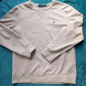 Beige sweatshirt från Polo Ralph Lauren - Säljer en stilren beige sweatshirt från Polo Ralph Lauren med klassisk rund hals och broderad logga på bröstet. Tröjan har långa ärmar och ribbade muddar vid ärmslut och nederkant. Perfekt för en clean och avslappnad look.