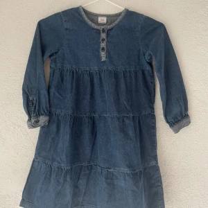 Jeansklänning Polarn O. Pyret 110/116 - Supersöt jeansklänning från Polarn O. Pyret i storlek 110/116. Klänningen har långa ärmar, knappar framtill och volangiga sömmar. Sann till storlek och oanvänd