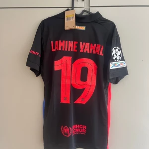 FC Barcelona Lamine Yamal Nike tröja - Snygg svart FC Barcelona fotbollströja från Nike med Lamine Yamal och nummer 19 på ryggen. Tröjan har röda och blå detaljer, officiella klubbmärken, sponsorloggor som Spotify och Ambilight tv samt UEFA Foundation-märke på ärmen. Tillverkad i lätt och ventilerande material.