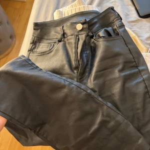 Svarta byxor i skinnimitation - Snygga svarta byxor i skinnimitation med klassisk femficksdesign och hög midja. Byxorna har raka ben och stängs med dragkedja och knapp. Perfekta för dig som vill ha en edgy look och gillar trendiga material.