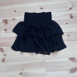 Svart volangkjol med resårmidja - Supersöt svart kortkjol med dubbla volanger och bred smock i midjan. Kjolen är luftig och har en flowy känsla, perfekt för att skapa en cool och trendig look. Passar lika bra till sneakers som boots.