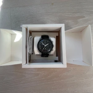 Omega x Swatch MoonSwatch svart klocka - Omega x Swatch MoonSwatch med svart urtavla och svart armband. Klockan har sportig design med tydliga visare och kronograf-funktion. Kommer i originalförpackning med manual och tillbehör. Perfekt för dig som gillar stilrena accessoarer. HELT NY FICK DEN I FÖDELSEDAGS PRESENT PAPPER FINNS SOM MAN SER PÅ BILDERNA 