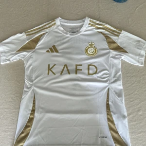 Al Nassr Ronaldo Tröja - Snygg o aldrig använd Al Nassr fotbollströja från Adidas i vitt med guldiga detaljer och tryck. Tröjan har Ronaldo 7 på ryggen, klubbmärke på bröstet och KAFD-logga framtill. Slim fit-modell i lätt och ventilerande polyestermaterial, perfekt för match eller träning.