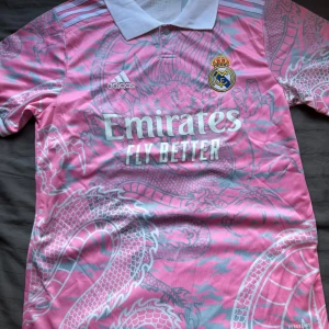 Rosa Real Madrid x Adidas fotbollströja - Snygg rosa Real Madrid fotbollströja från Adidas med vit krage och coolt drakmönster i ljusgrått. Tröjan har korta ärmar, klubbmärke på bröstet och 'Emirates Fly Better' tryckt framtill. Perfekt för dig som vill sticka ut på planen eller läktaren.