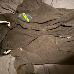 Svart AIK Nike hoodie med dragkedja - Svart hoodie från Nike med AIK-emblem på bröstet och klassisk swoosh-logga. Tröjan har huva med snörning, hel dragkedja och är tillverkad i mjukt material som passar perfekt för fotbollshäng eller chill. Snygg och sportig stil för dig som gillar AIK. Kan diskutera priset