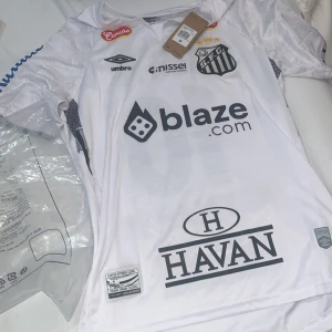 Santos FC Neymar Jr 10 matchtröja - Vit Santos FC fotbollströja från Umbro med Neymar Jr och nummer 10 på ryggen. Tröjan har flera tryckta sponsorer, klubbmärke på bröstet och korta ärmar. Tillverkad i lätt och ventilerande material, perfekt för dig som älskar fotboll. Helt ny strl M svår att få tag på. Pris kan diskuteras!