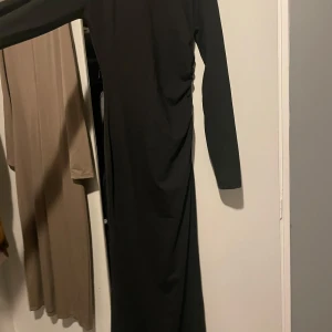  maxiklänning - Helt ny, aldrig använt. En svart långklänning med långa ärmar och enkel, stilren design. Klänningen har en lätt rynkning i sidan och är figurnära med en slits nedtill. Perfekt för dig som gillar minimalistisk och clean stil.
