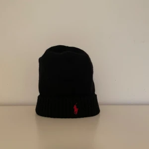 Svart mössa från Polo Ralph Lauren - Svart ribbstickad mössa från Polo Ralph Lauren med klassisk röd broderad logga framtill. Mössan har uppvikt kant och är tillverkad i mjukt material som håller dig varm. Perfekt accessoar för kyliga dagar och snygg till alla outfits.