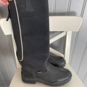 Svarta ridstövlar från Steeds - Snygga svarta ridstövlar från Steeds med högt skaft och robust sula. Tillverkade i syntetmaterial med diskreta reflexdetaljer längs sidan. Perfekta för dig som vill ha stilrena och funktionella stövlar till ridning.