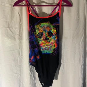 Svart baddräkt neonmönster - Cool svart baddräkt från Flipturns med färgglatt neonmönster i form av en döskalle framtill. Smala axelband i rosa och lila som korsas i ryggen. Perfekt för dig som vill sticka ut på stranden eller i poolen.