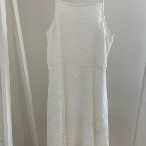 Vit spetsklänning med ärmlös design - Superfin vit klänning i spets med ärmlös modell och rund halsringning. Klänningen har en rak passform och är täckt av ett vackert blommigt spetsmönster. Perfekt för sommarens alla tillfällen och ger en fräsch och romantisk look.