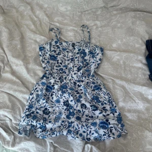 Blommig sommarklänning med smala axelband - Supersöt kort klänning med smala axelband och blått blommönster på vit botten. Perfekt för varma dagar, med en luftig passform och markerad midja. Materialet känns lätt och skönt mot huden.