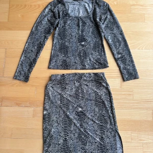 Glitzy kjol och blus med glittrig ormmönster - Kjol och blus från Glitzy i glittrigt ormmönster i svart och grått. Kjolen har en rak passform och en slits på sidan för extra edge. Materialet är stretchigt och glansigt, perfekt för dig som vill sticka ut.