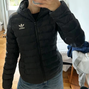 Svart pufferjacka från Adidas - Svart pufferjacka från Adidas med huva och dragkedja framtill. Jackan har Adidas logga på bröstet och är quiltad för extra värme. Tillverkad i syntetmaterial och har en clean, sportig look som funkar till jeans eller träningsbyxor.