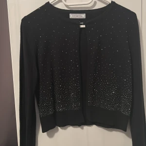 Svart kofta med glittriga detaljer Cosmoda - Svart kofta från Cosmoda med långa ärmar och glittriga paljettdetaljer framtill. Vissa av paljetter har blivit missfärgade men det syns inte väl, annars är den i bra skick! Köpt i Spanien 💕