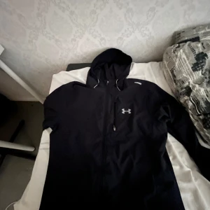 Svart vindjacka från Under Armour - Svart vindjacka från Under Armour med huva och dragkedja hela vägen. Jackan har en diskret logga på bröstet och justerbar dragsko i huvan. Tillverkad i lätt och smidigt syntetmaterial, perfekt för aktiva dagar utomhus. Snygg och sportig look.