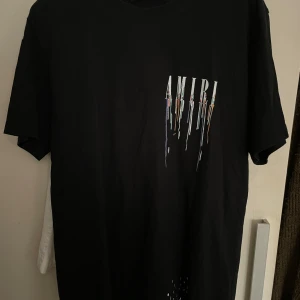 Svart AMIRI t-shirt med färgstänk - Svart t-shirt från AMIRI med stort tryck både fram och bak. Loggan är tryckt i vitt med färgglada droppar som ger en cool streetkänsla. Klassisk passform och rund hals. Perfekt för dig som gillar statement-plagg och vill sticka ut.