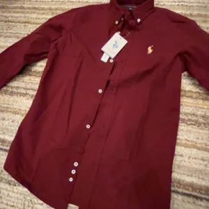 Vinröd skjorta från U.S. Polo Assn. - Snygg vinröd skjorta från Polo Ralph Louren. med klassisk krage och knappar framtill. Skjortan har lång ärm och ikonisk gul broderad logga på bröstet. Perfekt för dig som vill ha en stilren och tidlös look.