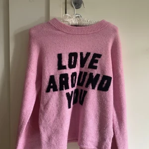 Rosa stickad tröja med text från H&M - Supermjuk rosa stickad tröja från H&M med svart texten 'LOVE AROUND YOU' framtill. Tröjan har rund halsringning och långa ärmar, perfekt för dig som gillar en chill och söt stil. Passar perfekt till jeans eller kjol för en trendig look.