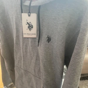 Grå hoodie från U.S. Polo Assn. - Snygg grå hoodie från U.S. Polo Assn. med svart logga broderad på bröstet och svarta snören i huvan. Tröjan är helt oanvänd som ni ser så finns polo prislappen kvar på tröjan och kvitto finns digitalt på polos hemsida, ta kontakt med mig för mer info.