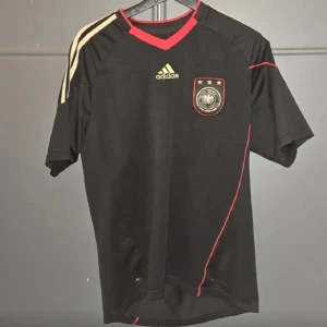 Svart Adidas Tyskland fotbollströja - Snygg svart fotbollströja från Adidas med Tysklands landslagsmärke på bröstet. Tröjan har korta ärmar, röda detaljer vid halsen och längs sidan samt guldiga Adidas-ränder på axlarna. Tillverkad i lätt och ventilerande material, perfekt för match eller träning.