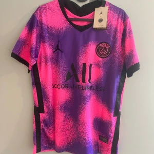 PSG Mbappé fotbollströja rosa/lila - Snygg PSG fotbollströja med Mbappé 7 på ryggen. Tröjan har ett unikt rosa och lila mönster, svarta detaljer och klubbmärke på bröstet. Tillverkad i lätt och ventilerande polyester, perfekt för match eller träning. Jordan-logga på bröstet och sponsortryck.