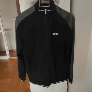 Svart vindjacka från BOSS - Svart vindjacka från BOSS med grå detaljer på axlar och ärmar. Jackan har hel dragkedja framtill, ståkrage och vit BOSS-logga på bröstet. Sportig look med ribbade muddar och smal passform. Men det finns några fläckar. Perfekt för dig som gillar stilren och enkel design.