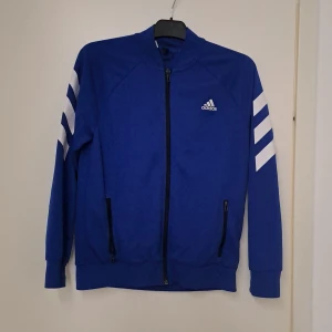 Blå Adidas vindjacka med dragkedja - Blå vindjacka från Adidas med vita ränder på ärmarna och klassisk logga på bröstet. Jackan har hel dragkedja framtill, två fickor med dragkedja även innerficka och ribbade muddar. Perfekt för sport eller chill, snygg och enkel att matcha.
